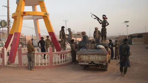 Des combattants en treillis autour d'un pick-up sur une route poussiéreuse au Mali. Un homme en contre-jour, debout sur le véhicule, brandit un fusil automatique.